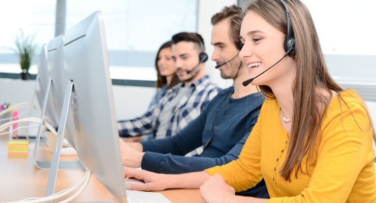 portrait of beautiful and cheerful young woman telephone operator with headset working on desktop computer in row in a customer service call support helpline business center with teamworker in background